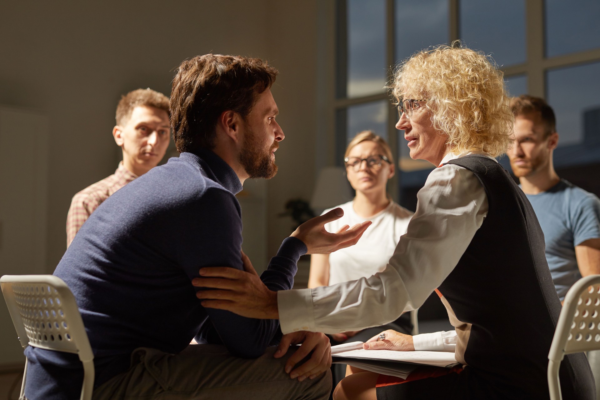 Serious sad bearded man sitting in circle of patients and sharing his problems at group therapy session, mature female psychiatrist touching his arm and supporting him